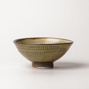 May include: A ceramic bowl with a tapered foot and a wide, open rim. The exterior features a repeating vertical line pattern etched into the olive-green glaze. The interior has a smooth, light-toned finish, while the base shows a dark brown tone.