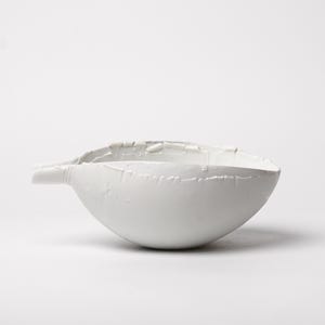 May include: A white ceramic bowl with a unique, textured surface and a small spout. The bowl has a rounded shape and is set against a plain white background. The design features visible seams and a rough texture.