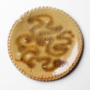 May include: A round ceramic plate with a scalloped edge. The plate is glazed in a light brown color with a brown and white abstract design.