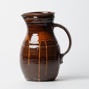 May include: A handcrafted brown ceramic pitcher with a glossy finish. The pitcher features a rounded body, a curved handle, and a spout for pouring. The glaze has a dripped effect, adding a unique touch to this rustic kitchenware.