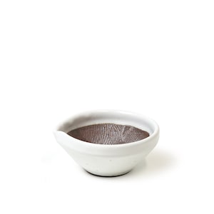 May include: A small, white ceramic bowl with a textured, brown interior. The bowl has a slightly irregular shape with a pouring spout on one side. The exterior is smooth and glazed, while the interior features a ridged pattern.
