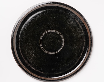 May include: A round, black ceramic plate with a slightly raised rim and a recessed center. The plate has a glossy finish and a dark, textured surface. The rim has a brown accent. The plate is suitable for serving food.