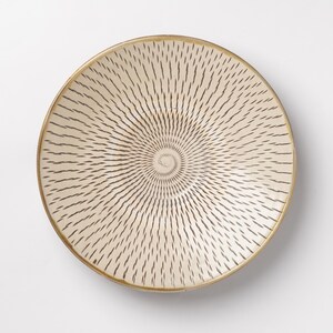 May include: A round ceramic bowl with a cream-colored base and a dark brown radial pattern of small, dash-like marks that spiral outward from the center toward the rim. The bowl features a thin, solid brown line along the circular edge.