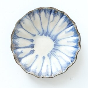 Puede incluir: Un cuenco pequeño y redondo de cerámica con borde festoneado. El cuenco presenta un interior blanco con un patrón radial azul que emana del centro. El borde es de color marrón oscuro y texturizado. El cuenco se utiliza probablemente para servir alimentos o como pieza decorativa.