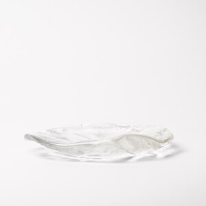 May include: A clear, decorative glass dish with a wavy, organic shape. The dish is transparent, allowing light to pass through, and sits on a white surface. The design is reminiscent of a leaf or a natural form.