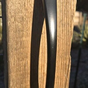 May include: A matte black metal door handle with a smooth, elongated, and slightly curved design. The handle features a pointed top and a rounded bottom, mounted vertically against a textured, light brown wooden surface with visible grain.