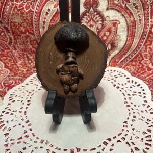 Puede incluir: Una pequeña escultura de madera de un personaje de hongo con un cuerpo marrón y un sombrero de hongo negro. La escultura está montada en una rodaja de madera y se muestra en un encaje blanco.