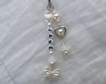 May include: A beaded charm with the name "JANE" spelled out in white letter beads. The charm features pearl and heart-shaped accents, a bow charm, and a black cord loop for attachment. The color palette includes white, silver, and pink.