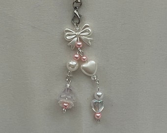 May include: A delicate phone charm featuring a white bow, pearl beads, and pink heart and flower accents. The charm is attached to a black cord and silver-toned hardware, creating a charming accessory.