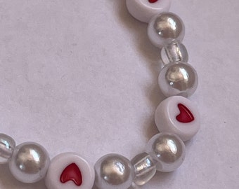 May include: A white pearl bracelet with red heart charms. The bracelet is made of small white pearls and has three red heart charms.