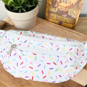 May include: White fanny pack with a colorful sprinkle pattern. The fanny pack has two zippered compartments and a white adjustable strap.