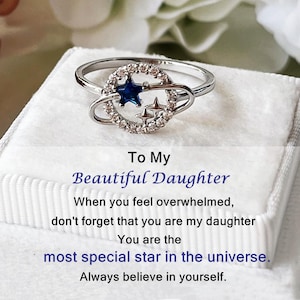 May include: A silver ring with a celestial design. The ring features a blue star and smaller stars, surrounded by clear stones. The text "To My Beautiful Daughter" is visible.