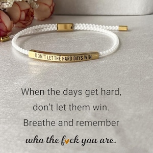 Könnte beinhalten: Weißes geflochtenes Armband mit goldfarbenen Akzenten. Das Armband hat einen goldenen Balken mit der Aufschrift "DON'T LET THE HARD DAYS WIN". Das Bild enthält auch den Text: "When the days get hard, don't let them win. Breathe and remember who the f*ck you are."