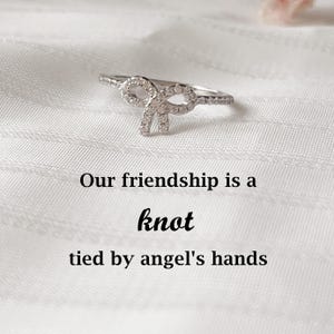 Könnte beinhalten: Ein silberner Ring mit Schleifendesign, besetzt mit kleinen, klaren Edelsteinen. Der Ring liegt auf einer weißen, strukturierten Oberfläche. Der Text auf dem Bild lautet: "Our friendship is a knot tied by angel's hands."