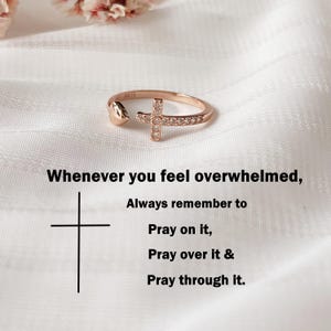 Op de afbeelding: Roségouden ring met een kruisontwerp, versierd met kleine, heldere stenen. De ring staat tegen een witte, getextureerde achtergrond. Tekst: "Wanneer je je overweldigd voelt, denk er dan altijd aan om erover te bidden, erover te bidden en erdoorheen te bidden."