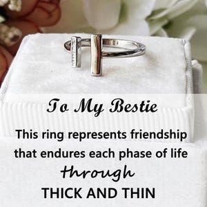 May include: A silver ring with a vertical bar and a smaller bar with embedded crystals. The ring is displayed on a white surface with the text "To My Bestie" and "This ring represents friendship that endures each phase of life through thick and thin."