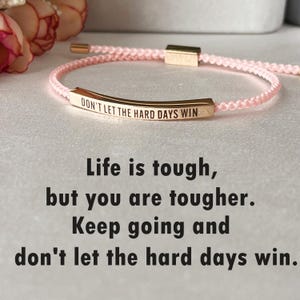 Puede incluir: Un brazalete de cordón trenzado rosa con un dije de barra dorada que dice "DON'T LET THE HARD DAYS WIN". El brazalete es un recordatorio para mantenerse positivo y seguir adelante.