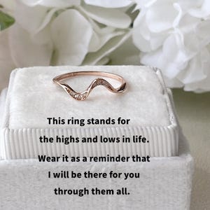 May include: Rose gold ring with a wave design, set with small, clear stones. The ring is displayed on a white, textured surface. Text on the image reads: "This ring stands for the highs and lows in life."