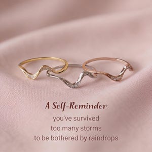 May include: Three wave-shaped rings in gold, silver, and rose gold tones, each featuring small embedded stones. The text reads: A Self-Reminder, you've survived too many storms to be bothered by raindrops. The rings are arranged in a horizontal row.