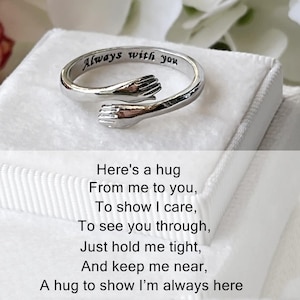 May include: Silver-colored ring with an embracing hands design. The ring has the words "Always with you" engraved on the band. The image also includes a poem about hugs.