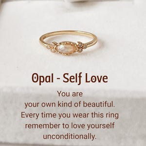 May include: A gold ring with an oval opal stone and small accent stones on each side. The ring is set against a white background. Text on the image reads "Opal - Self Love".