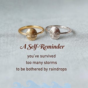 May include: Two rings, one gold-toned and one silver-toned, each featuring a small oval face engraved with a wave pattern. The text below reads: A Self-Reminder, you've survived too many storms to be bothered by raindrops. The rings are thin bands.
