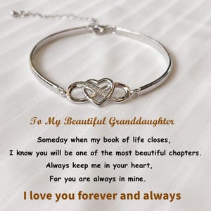 May include: A silver bracelet with a heart and infinity symbol design. The bracelet has a delicate chain and a heart-shaped centerpiece adorned with small, sparkling stones. Text on the background reads "To My Beautiful Granddaughter."
