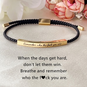 Peut inclure: Bracelet tressé noir avec des accents dorés et une barre dorée gravée du texte "Remember who the f*ck you are." Le bracelet contient également le texte "When the days get hard, don't let them win. Breathe and remember who the f*ck you are."