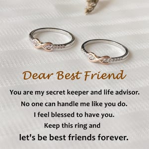 May include: Two silver rings with an infinity symbol design, accented with rose gold and small clear stones. The rings are on a white surface with the text "Dear Best Friend" and a friendship message.