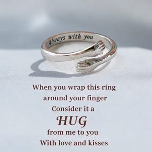 Puede incluir: Un anillo abierto de tono plateado con dos manos que se encuentran en el centro formando un abrazo. El interior de la banda está grabado con el texto Always with you. Debajo del anillo, el texto dice: When you wrap this ring around your finger Consider it a HUG from me to you With love and kisses.