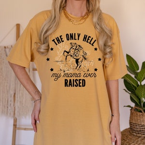 Könnte beinhalten: Ein senfgelbes T-Shirt mit einer schwarzen Grafik eines sich aufbäumenden Pferdes. Die Grafik ist von Sternen umgeben und dem Text "THE ONLY HELL MY MAMA EVER RAISED".