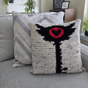 May include: Two decorative cushions on a grey sofa. One cushion is cream-coloured with a black and red graphic design. The other cushion has a grey and white chevron pattern. The cushions add a decorative touch to the living room.