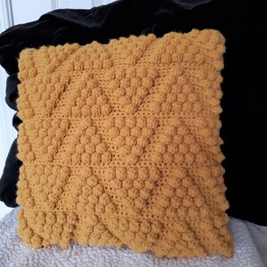 May include: A square yellow crocheted pillow with a textured, bumpy surface. The pillow is sitting on a white surface.