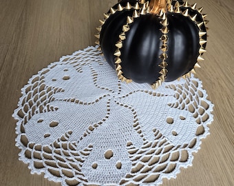 Könnte beinhalten: Ein mattschwarzer Kürbis, verziert mit Reihen goldener Metallspitzen und einem goldenen Stiel, ruht auf einer runden, weißen Häkeldeckchen mit einem sich wiederholenden Muster aus Geisterformen und durchbrochenen Details am gewellten Rand.