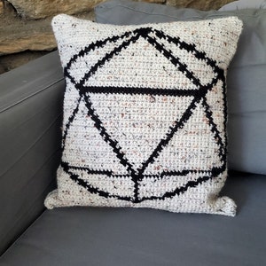 May include: A square, crocheted pillow with a geometric black design on a speckled white background. The pillow is resting on a gray surface, with a stone wall in the background. The pillow is approximately 16 inches square.