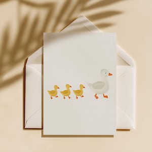 May include: A greeting card featuring a white goose leading three yellow ducklings. The illustration is on a white card, set inside a white envelope, with a neutral background and leaf shadow.