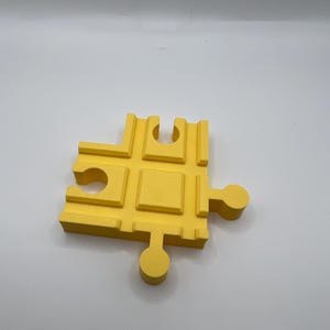 May include: A bright yellow puzzle piece with a unique, geometric design. The piece features a series of rectangular and circular cutouts, and a circular peg extending from the side. The piece is made of a solid material and is set against a white background.