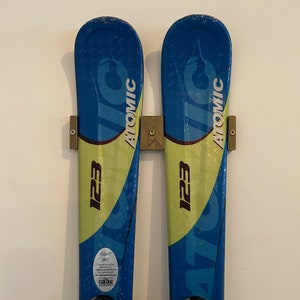 May include: Pair of blue and green Atomic 123 skis. The skis are mounted on a wall with a brown ski holder. The skis have the word "Atomic" printed on them. The skis have a blue and green design.