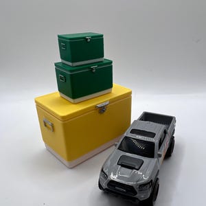 K&ouml;nnte beinhalten: Ein Set aus gelben, gr&uuml;nen und wei&szlig;en K&uuml;hlboxen mit einem grauen Toyota Tacoma Spielzeug-Truck. Die K&uuml;hlboxen sind gestapelt, mit einer gro&szlig;en gelben K&uuml;hlbox unten und zwei gr&uuml;nen K&uuml;hlboxen dar&uuml;ber.