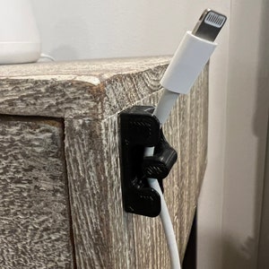 May include: A black cable management clip attached to a wooden surface, holding a white charging cable with a lightning connector.