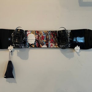 May include: A black snowboard with black bindings and a colorful graphic design is mounted on a white wall. The snowboard is held in place by white wall mounts. A black bag hangs from one of the mounts.