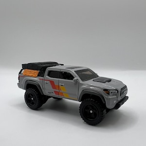 May include: A gray toy truck with black wheels and a black roof rack. The truck has orange, yellow, and red stripes on the side. The truck is a miniature replica of a real vehicle.