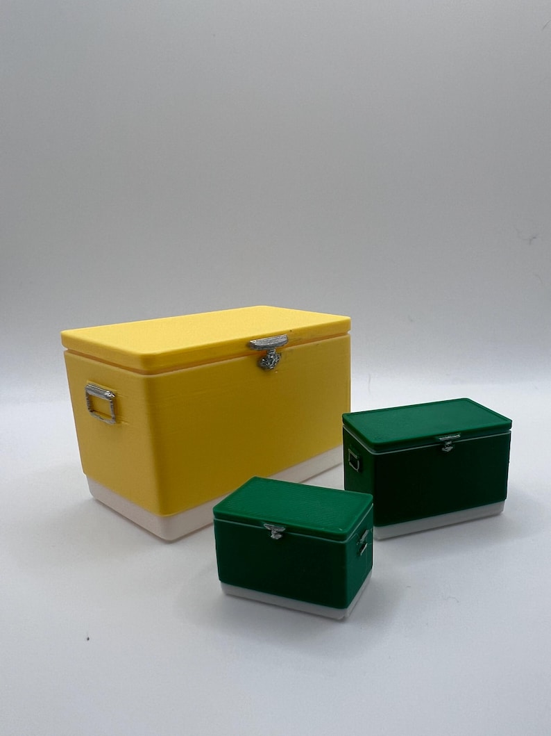 K&ouml;nnte beinhalten: Drei rechteckige K&uuml;hlboxen in verschiedenen Gr&ouml;&szlig;en. Die gr&ouml;&szlig;te ist gelb mit wei&szlig;em Sockel, die mittleren und kleinen K&uuml;hlboxen sind gr&uuml;n mit wei&szlig;en Sockeln. Alle haben silberne Verschl&uuml;sse und Griffe.