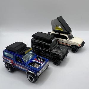 May include: Three toy off-road vehicles. A blue Nissan Patrol with red and white accents, a black Land Rover Defender, and a white SUV with a pop-up tent. All have black wheels and roof racks.