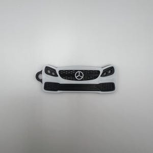 May include: A white and black 3D-printed car front grill with a key ring attachment. The design features a central emblem, headlights, and a textured lower grill. The object is a miniature replica of a car's front.