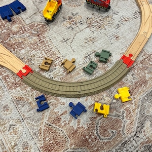 May include: A wooden train track set with a red train engine, a yellow dump truck, and various track pieces. The track pieces are in different colors, including blue, yellow, and green. The track is laid out on a patterned rug.