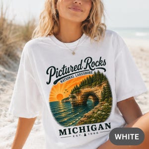 Può includere: Una t-shirt bianca con una stampa grafica di Pictured Rocks National Lakeshore in Michigan. Il design include un tramonto sull'acqua, un arco di roccia naturale e pini, con il testo Pictured Rocks National Lakeshore Michigan Est. 1966.