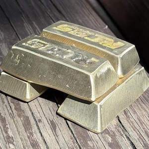 May include: Three rectangular gold-colored metal bars stacked in a pyramid formation. Each bar features a brushed finish and the word "GOLD" embossed in capital letters on the top surface. The metal has a metallic sheen with visible texture.
