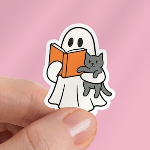 May include: A white ghost sticker with black eyes and an orange book. The ghost is holding a gray cat. The sticker is held in a hand against a pink background.