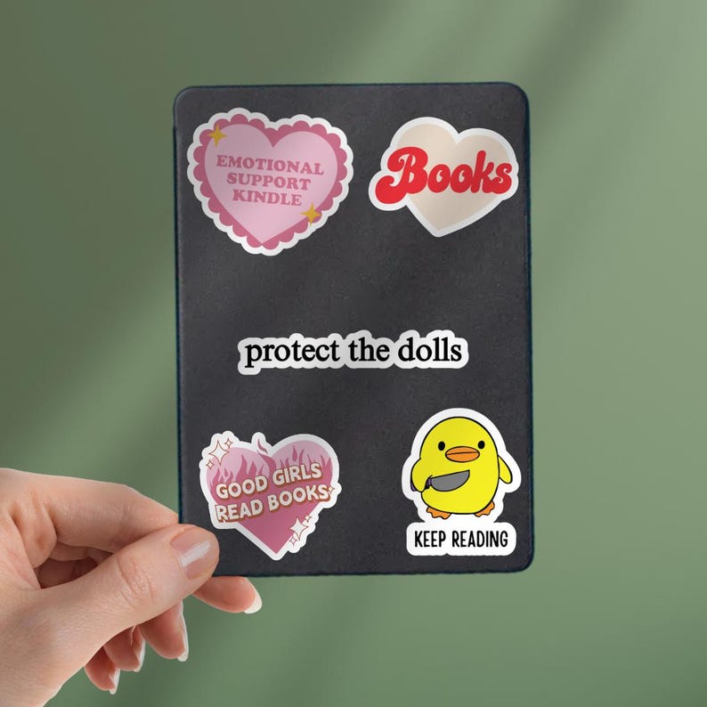 May include: A collection of stickers on a black background. The stickers include a pink heart with the text "EMOTIONAL SUPPORT KINDLE", a heart-shaped sticker with the word "Books", and a yellow chick sticker with a knife. The text "protect the dolls" is also visible.